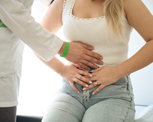 Cropped photo of female patient with stomachache seated on exam table while doctor palpating her abdomen