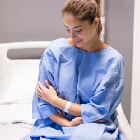 Young female patient in hospital ward,dressed in blue gown,sitting on bed and waiting to be examined by doctor,smiling softly and satisfied with service and services provided,individual hospital room